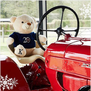 Kingsland Teddybär Asaan sitzt auf rotem Porsche-Traktor, trägt blauen Pullover mit 20-Jahre-Logo – perfektes Geschenk für Reiterfans.